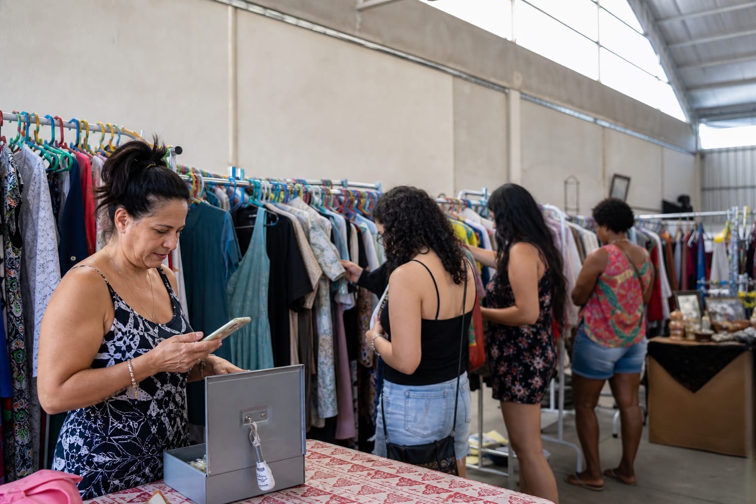 Bazar de brechó em galpão com araras cheias e expositora atendendo no balcão